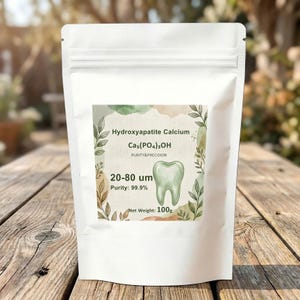 May include: A white pouch with a label displaying text: "Hydroxyapatite Calcium, Cas(PO4)3OH, Purity & Precision, 20-80 um, Purity: 99.9%, Net Weight: 100g." The label includes a tooth graphic. The pouch is on a wooden surface.
