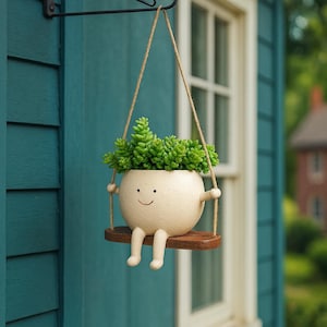 May include: A whimsical hanging planter featuring a smiling, off-white ceramic pot with arms and legs, suspended from a wooden swing. The pot is filled with vibrant green succulents. The planter hangs from a black metal bracket on a teal wall.
