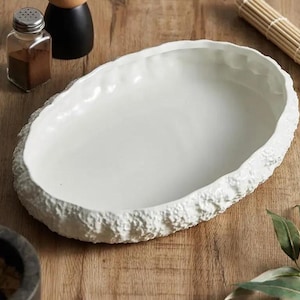 May include: A white, oval serving dish with a textured, stone-like rim. The dish is empty and sits on a wooden surface, alongside a small glass jar with a metal lid, a black pepper grinder, and a bamboo sushi mat.