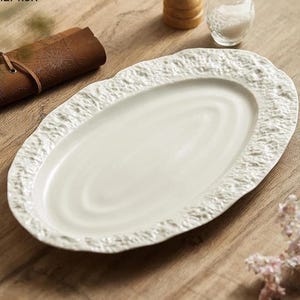 May include: An oval, off-white serving platter with a textured, raised border. The centre of the platter is smooth and slightly concave. The platter is on a wooden surface, suggesting a rustic or farmhouse style.