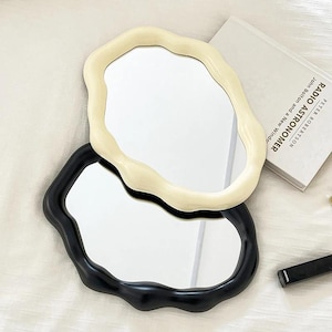 May include: Two decorative mirrors with irregular, wavy frames. One mirror has a cream-colored frame, and the other has a black frame. The mirrors are placed on a white surface, with a book and a black object visible in the background.