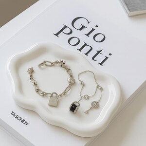 May include: A white, cloud-shaped ceramic tray holds two pieces of jewelry. A silver chain bracelet with a padlock charm and a silver necklace with a key pendant. The tray rests on a book with the title "Gio Ponti".