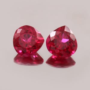Könnte beinhalten: Zwei lose birnenförmige Edelsteine mit einer leuchtend tiefroten Farbe und facettierten Oberflächen. Die Steine weisen eine hohe Klarheit und Brillanz auf und reflektieren das Licht durch ihre präzise geschliffenen Facetten.