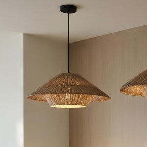 May include: Two woven pendant lights in a natural, light brown hue hang from the ceiling. The lamps have a layered, conical design, with the light fixtures visible inside. The lamps are above a beige sofa in a living room.