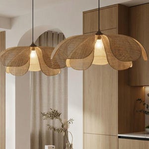 Puede incluir: Dos lámparas colgantes con pantallas tejidas en forma de pétalo cuelgan sobre una encimera de mármol. La cocina cuenta con gabinetes de madera clara, una mesa ovalada blanca y dos sillas de madera. Un jarrón con vegetación y un cuenco de peras están en la encimera.