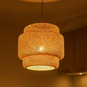 May include: A woven rattan pendant light fitting with a layered, cylindrical design. The light fitting hangs from the ceiling, illuminating a kitchen setting. A teapot and teacups sit on a wooden table below.