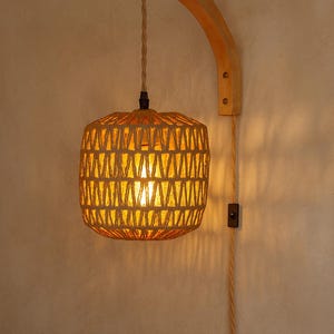 Może przedstawiać: Tkana lampa wisząca z rattanu o ciepłym blasku. Lampa wisi na zakrzywionym drewnianym ramieniu przymocowanym do ściany. Projekt ma kwadratowy kształt z geometrycznym wzorem i włącznikiem/wyłącznikiem.