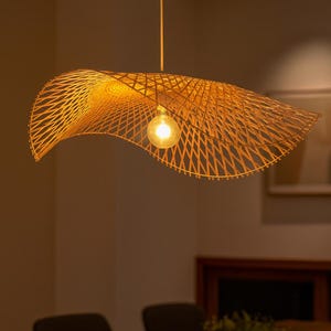Może przedstawiać: Tkana bambusowa lampa wisząca o unikalnym, kapeluszowatym kształcie. Lampa jest zawieszona pod sufitem i posiada pojedynczą, odsłoniętą żarówkę. Ciepłe, naturalne odcienie bambusa tworzą przytulną atmosferę. Średnica lampy wynosi około 76 cm.