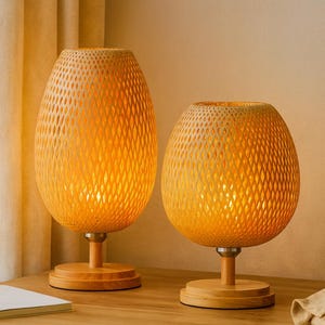 May include: Two decorative table lamps with woven, egg-shaped shades. The shades are a warm yellow color and sit atop wooden bases. The lamps are lit, emitting a soft, inviting glow. The lamps are on a wooden table.