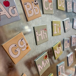 May include: Alphabet magnets with uppercase and lowercase letters, each with a tracing guide. The letters are on colorful backgrounds with illustrations like a soccer ball, apple, and giraffe. The magnets are arranged on a metallic surface.