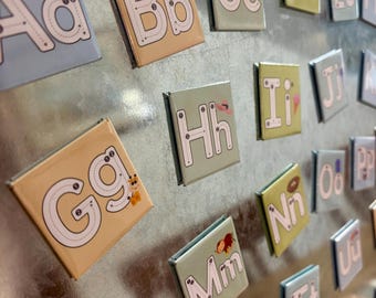 May include: Alphabet magnets with uppercase and lowercase letters, each with a tracing guide. The letters are on colorful backgrounds with illustrations like a soccer ball, apple, and giraffe. The magnets are arranged on a metallic surface.