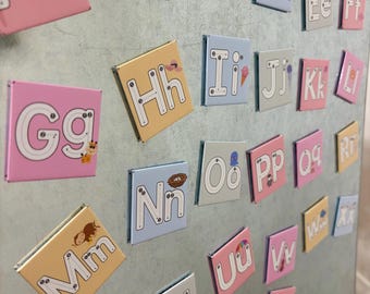 May include: Alphabet magnets in various pastel colors, including pink, yellow, and blue. Each magnet features a letter of the alphabet with a corresponding illustration, such as an apple for 'A' and a soccer ball for 'B'. The magnets are arranged on a metallic surface.