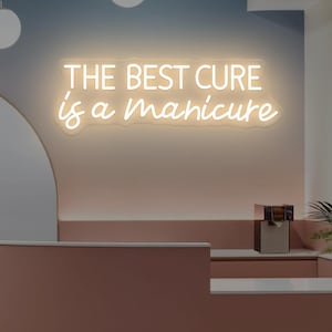 Könnte beinhalten: Ein Neonschild mit der Aufschrift "THE BEST CURE is a manicure" in einem Salon. Das Schild leuchtet in warmem Licht. Der Salon hat einen geschwungenen, staubrosa Empfangstresen und eine neutrale Farbpalette.