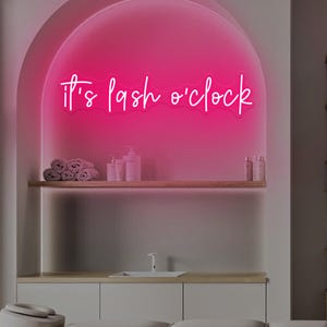 Puede incluir: Un letrero de neón rosa con el texto "It's lash o'clock". El letrero está montado en una pared con una alcoba curva. Debajo, se ve una camilla de masaje y otros equipos de salón.
