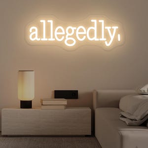 以下が含まれることがあります： 白い筆記体で「allegedly」と書かれたネオンサイン。ベージュの壁に設置され、ランプと本が置かれたナイトテーブルの上にあります。部屋はニュートラルな色使いです。
