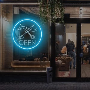 May include: A storefront with a neon blue "OPEN" sign featuring scissors and hair. The sign is in a circular frame. The shop's window displays the interior, including a seating area and people. The words "Booking" and "Walk in" are visible.