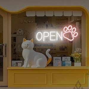以下が含まれることがあります： 黄色いファサードと白い猫の像がある店の正面。ネオンサインには足跡とともに「OPEN」と書かれています。猫はオレンジ色の尻尾と耳をしています。ショーウィンドウには様々な商品が展示されています。