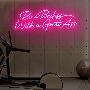 May include: A vibrant pink neon sign with the phrase "Be a Badass With a Great Ass" in a cursive font. The sign is mounted on a neutral-toned wall in a gym setting, with exercise equipment visible in the foreground.