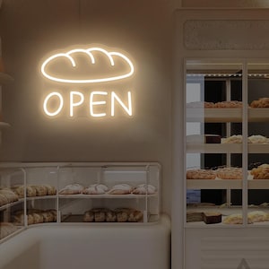 May include: A bakery interior featuring a neon sign with a bread loaf design and the word "OPEN". Display cases showcase various baked goods. The overall color scheme is neutral, with white and beige tones.