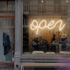 May include: A shop window features a neon sign in cursive that reads "open". The interior is visible, with a bench and pillows in front. The building's exterior is stone and brick. The shop is open for business.