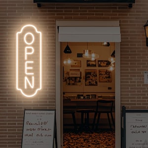 May include: A glowing neon "OPEN" sign in a vertical, rectangular shape is mounted on a brick wall. The sign is a warm, off-white color and is positioned to the left of a storefront entrance. The entrance has a dark awning and a vintage-style lamp.