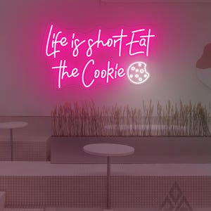 May include: A bright pink neon sign with the phrase "Life is short Eat the Cookie" in a handwritten font. The cookie is represented by a white outline with a few small circles. The sign is mounted on a white wall.