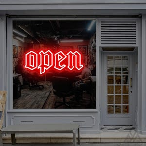 May include: A shopfront window featuring a bright red neon sign that displays the word open in a gothic-style font. The window provides a view into an interior space with chairs and artwork, while a grey door sits to the right of the glass.