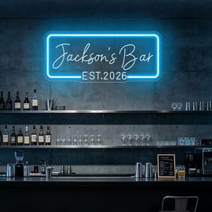 Puede incluir: Un letrero de neón rectangular con un brillo azul intenso que muestra el texto Jackson's Bar y EST. 2026 en una fuente cursiva y sans-serif. El letrero está montado en una pared de hormigón gris oscuro sobre estantes llenos de botellas y cristalería.