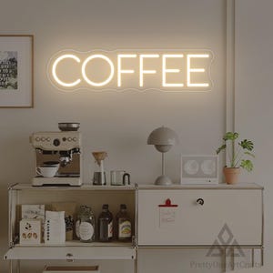 Puede incluir: Un letrero de neón con la palabra "COFFEE" en blanco cálido. Debajo, una estación de café con una máquina de espresso, granos de café y una pequeña planta en maceta, creando un rincón de café acogedor.