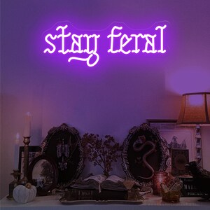 May include: A neon sign in purple script reads "stay feral" above a shelf with gothic decor. The shelf displays framed art, a candle, a pumpkin, and an open book, creating a spooky aesthetic.