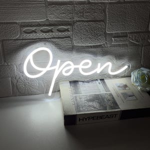 Op de afbeelding: Een wit neon "Open" bord in een cursief lettertype wordt verlicht tegen een getextureerde witte bakstenen muur. Een boek met het woord "HYPEBEAST" is zichtbaar. Een witte roos en een boek liggen op het oppervlak.