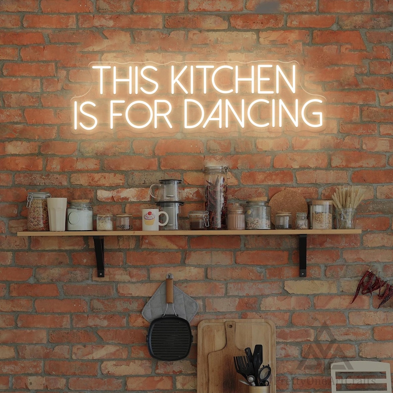 May include: A neon sign with the text "THIS KITCHEN IS FOR DANCING" is mounted on a red brick wall. Below the sign, a wooden shelf holds various kitchen items, including jars, mugs, and utensils. A black grill pan hangs below.