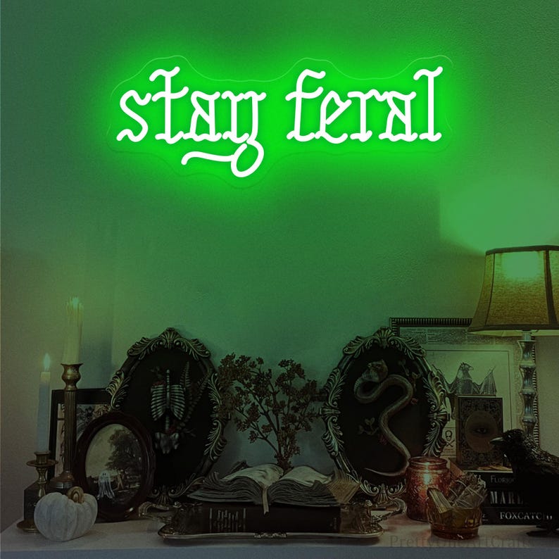 May include: A neon green sign displaying the words "stay feral" in a decorative font. The sign is mounted on a green wall above a shelf with framed pictures, candles, and decorative objects, creating a gothic or Halloween theme.