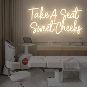 Può includere: Un interno di una spa con un lettino bianco, attrezzature mediche e un'insegna al neon con la scritta "Take A Seat Sweet Cheeks". La stanza ha una palette di colori neutri con pareti beige e mobili chiari. Un piccolo computer è su un supporto.