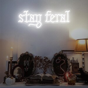 May include: A white neon sign that reads "stay feral" in a gothic font is mounted on a white wall. Below the sign is a shelf with framed pictures, books, candles, and decorative objects, creating a gothic or witchy aesthetic.