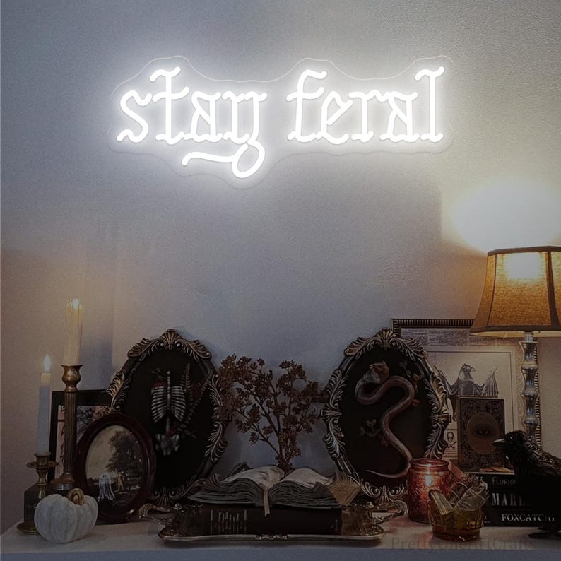 May include: A white neon sign that reads "stay feral" in a gothic font is mounted on a white wall. Below the sign is a shelf with framed pictures, books, candles, and decorative objects, creating a gothic or witchy aesthetic.