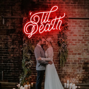 May include: A red neon sign that reads "'Til Death" in cursive script is mounted on a brick wall. A couple is embracing in front of the sign. The woman is wearing a white wedding dress, and the man is wearing a gray suit.