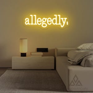 以下が含まれることがあります： 寝室のネオンサインは、明るい黄色で「allegedly」という単語を表示しています。サインは、ランプ、本、ベッドのあるナイトテーブルの上にある、明るいベージュの壁に取り付けられています。