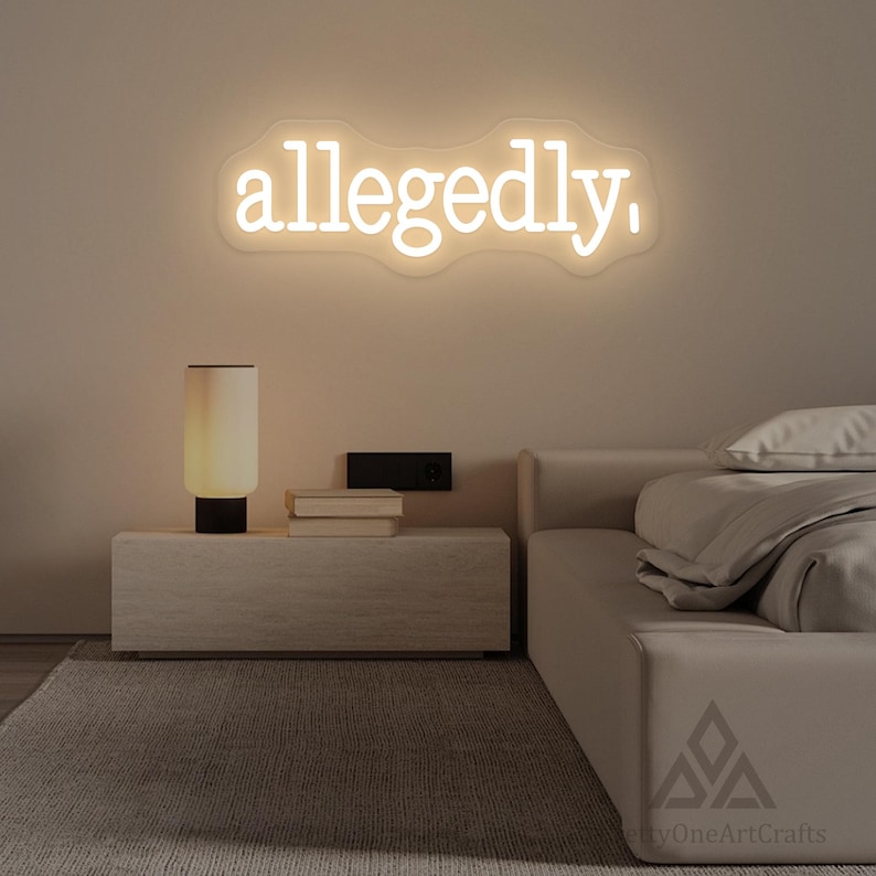 以下が含まれることがあります： 白い筆記体で「allegedly」と書かれたネオンサイン。ベージュの壁に設置され、ランプと本が置かれたナイトテーブルの上にあります。部屋はニュートラルな色使いです。