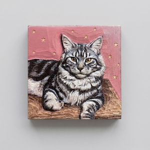 May include: A wooden carving of a cat with gray and black stripes, lying on a brown surface. The background is pink with gold star accents. The cat has yellow eyes and a fluffy chest.