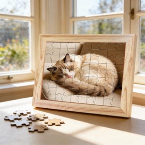 Puede incluir: Un rompecabezas enmarcado en madera con un gato durmiendo de pelaje marrón y blanco. El rompecabezas está parcialmente montado, con algunas piezas esparcidas sobre la mesa. El gato está acurrucado sobre un cojín beige.