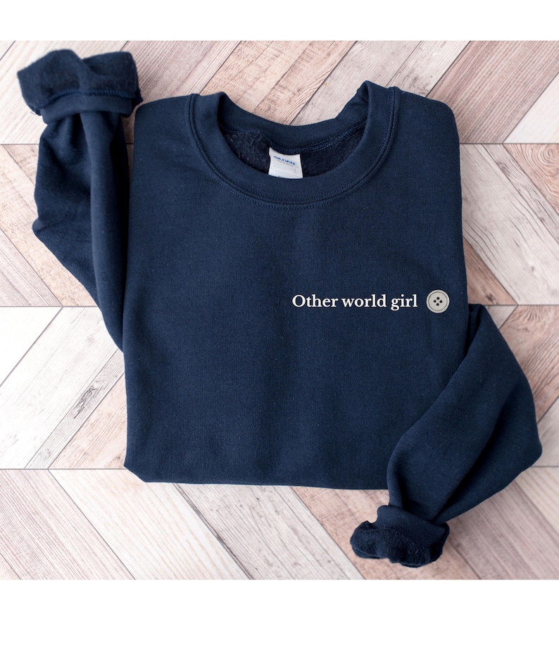 May include: A navy blue sweatshirt with the text "Other world girl" and a button graphic. The long-sleeved top has a crew neck and cuffed sleeves. The sweatshirt is displayed on a light wood-grain surface.