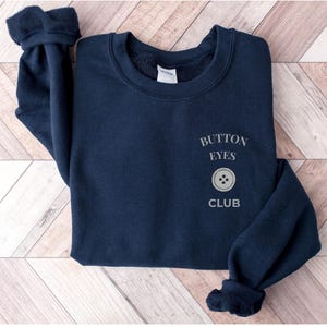 May include: A navy blue long-sleeve sweatshirt featuring the text "BUTTON EYES CLUB" printed in a light gray font on the left chest area, accompanied by a graphic of a four-hole button positioned between the words "EYES" and "CLUB".