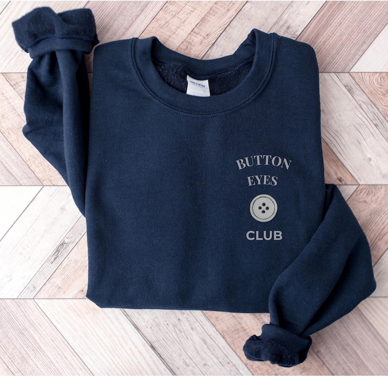 May include: A navy blue long-sleeve sweatshirt featuring the text "BUTTON EYES CLUB" printed in a light gray font on the left chest area, accompanied by a graphic of a four-hole button positioned between the words "EYES" and "CLUB".