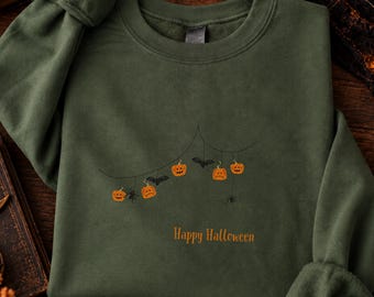 May include: An olive green crewneck sweatshirt featuring a central graphic of a thin black line draped like a string with hanging orange jack-o'-lanterns, black bats, and spiders. The text "Happy Halloween" is printed in orange below the graphic.