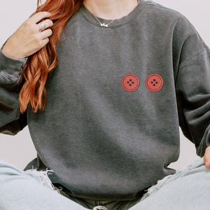 Puede incluir: Sudadera gris oscuro con dos gr&aacute;ficos de botones rojos. La sudadera tiene un corte relajado y cuello redondo. Los botones son de color rojo brillante y est&aacute;n centrados en el pecho.
