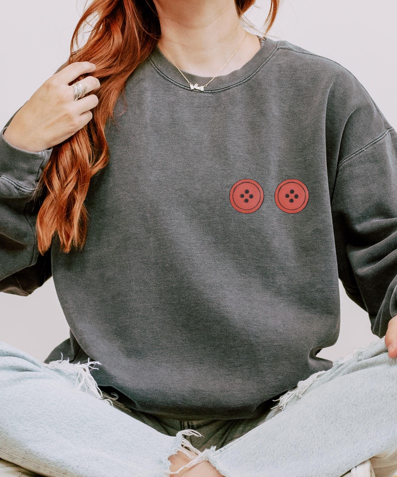 Puede incluir: Sudadera gris oscuro con dos gr&aacute;ficos de botones rojos. La sudadera tiene un corte relajado y cuello redondo. Los botones son de color rojo brillante y est&aacute;n centrados en el pecho.