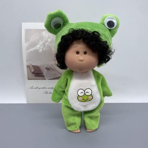 Può includere: Una bambola che indossa un costume da rana verde con un cappello abbinato. La bambola ha capelli ricci neri, un viso color pesca e occhi marroni. Il costume da rana ha una pancia bianca con una faccia da rana dei cartoni animati.