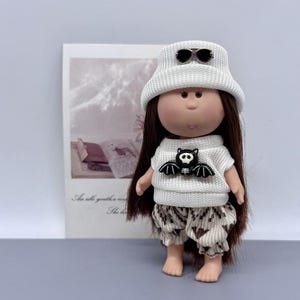 Può includere: Bambola con lunghi capelli castani, indossa un cappello di lana bianco con occhiali da sole, un maglione bianco con un disegno di pipistrello e pantaloncini fantasia. La bambola è davanti a un'immagine con testo.