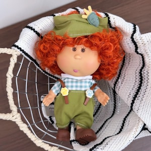 Può includere: Bambola in stile vintage con capelli ricci rosso vivo, indossa un cappello verde con un dettaglio giallo, una camicia a quadri blu e bianca, una salopette verde e scarpe marroni. La bambola è in un cestino di filo metallico con fodera a righe bianche e nere.